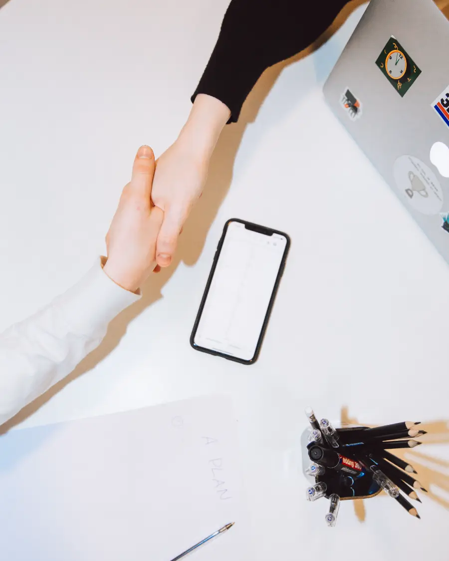 Two people shaking hands across a desk, beside a phone, a notebook and a pen.
