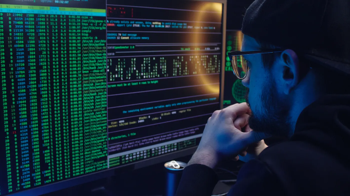 An engineer in a darkened operations room watching a live process table — running tasks, load average and an uptime counter — on one of two monitors.